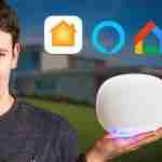 Man holding a white smart speaker (like a HomePod) in front of a modern house, with smart‑home app icons above.